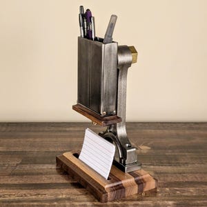 Può includere: Un'esclusiva organizzatore da scrivania in metallo e legno. Presenta un portapenne, un portablocco e un accento in ottone. L'organizzatore contiene penne e un blocco note con carta a righe. La base è in legno scuro.