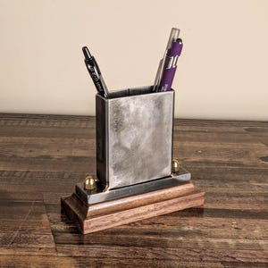 May include: A rectangular metal pen holder with a wooden base. The pen holder has a raw metal finish and holds several pens. The base is made of dark stained wood. Two brass-colored accents are on the front of the pen holder.
