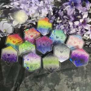 May include: A set of ten iridescent holographic dice with a heart symbol on each face. The dice are shaped like a 20-sided die and are arranged in a circle on a black surface.