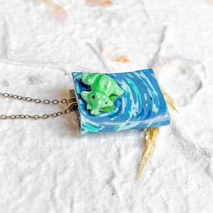 May include: A rectangular pendant necklace featuring a small, light green sculpted creature resting on a blue, water-patterned surface. The pendant is attached to a thin, dark bronze-toned metal chain, creating a detailed and colorful accessory.