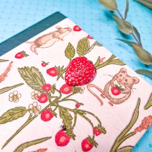 May include: A bright red strawberry sits atop a notebook with a whimsical strawberry and mouse print. The notebook has a green border and is set against a light blue background with eucalyptus leaves.