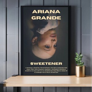 Puede incluir: Un póster con un retrato de Ariana Grande con el título "Sweetener" y una lista de canciones del álbum. El fondo es negro y el texto es de color dorado.