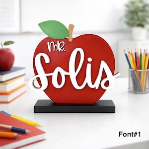 May include: A red apple-shaped desk sign with the name "Mr. Solis" in white script. The sign has a green leaf and a brown stem, and sits on a black base. The text "Font #1" is in the bottom right corner.
