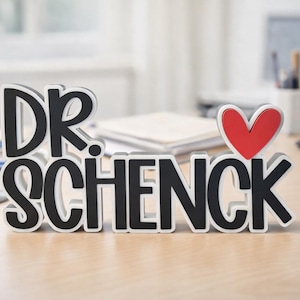 Puede incluir: Un letrero independiente que presenta el texto negro DR. SCHENCK con un s&iacute;mbolo de coraz&oacute;n rojo colocado sobre la letra K. Las letras tienen un borde blanco y un contorno negro grueso, creando un efecto dimensional en la superficie.