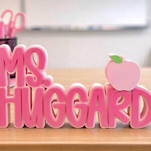 Puede incluir: Un letrero de escritorio rosa en 3D con el texto "MS. HUGGARD" en una fuente gruesa y superpuesta. El letrero incluye un peque&ntilde;o icono de manzana rosa claro con una hoja verde colocada sobre la letra D, creando una placa decorativa de escritorio.