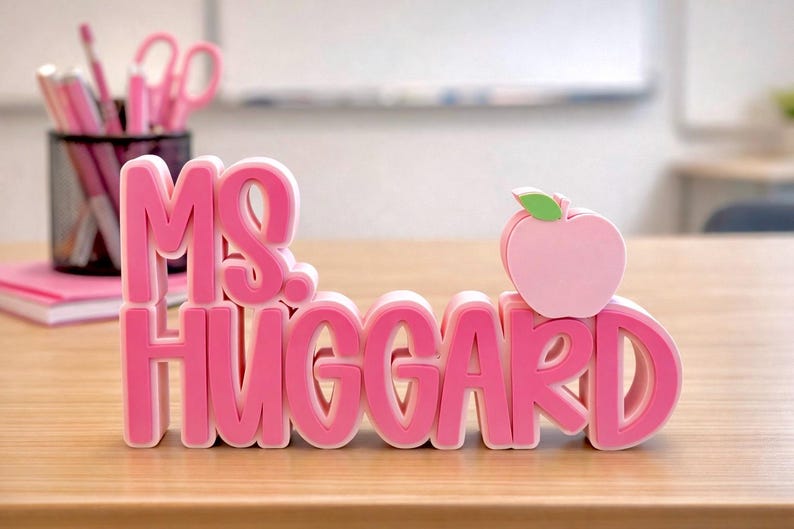 Puede incluir: Un letrero de escritorio rosa en 3D con el texto "MS. HUGGARD" en una fuente gruesa y superpuesta. El letrero incluye un peque&ntilde;o icono de manzana rosa claro con una hoja verde colocada sobre la letra D, creando una placa decorativa de escritorio.