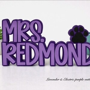 Puede incluir: Un letrero tridimensional con el texto MRS. REDMOND en letras color lavanda con un contorno morado oscuro. Un icono de huella negra est&aacute; colocado a la derecha del texto. El letrero est&aacute; construido con materiales en capas y se mantiene en pie.