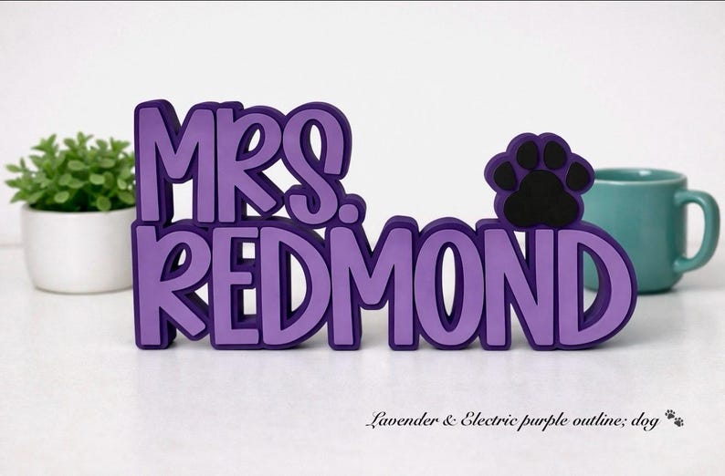 Puede incluir: Un letrero tridimensional con el texto MRS. REDMOND en letras color lavanda con un contorno morado oscuro. Un icono de huella negra est&aacute; colocado a la derecha del texto. El letrero est&aacute; construido con materiales en capas y se mantiene en pie.