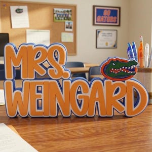 Puede incluir: Un letrero de escritorio en 3D con el texto MRS. WEINGARD en letras naranjas con bordes blancos y azules. Una ilustraci&oacute;n de una cabeza de caim&aacute;n verde con la boca abierta se sit&uacute;a a la derecha del nombre. El letrero est&aacute; hecho de capas.