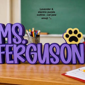 Puede incluir: Un letrero de escritorio personalizado con el nombre MS. FERGUSON en letras superpuestas de color lavanda y morado el&eacute;ctrico. El letrero incluye un icono de emoji de pata de gato amarillo y negro a la derecha. El texto tiene una fuente redondeada.