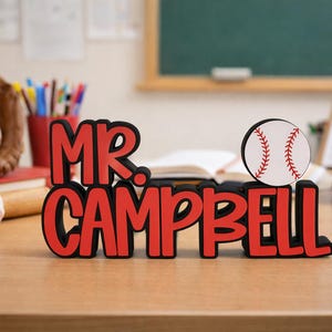 Puede incluir: Un letrero de escritorio personalizado con el texto MR. CAMPBELL en letras rojas en negrita con un borde negro. Un gr&aacute;fico circular de una pelota de b&eacute;isbol blanca con costuras rojas est&aacute; unido al lado derecho del letrero.