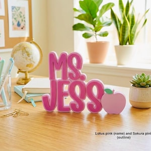 Puede incluir: Un letrero de escritorio personalizado con el nombre MS. JESS en letras rosas loto en relieve y negrita con un contorno rosa sakura, acompa&ntilde;ado de un peque&ntilde;o icono con forma de manzana rosa claro unido al lado derecho del nombre.