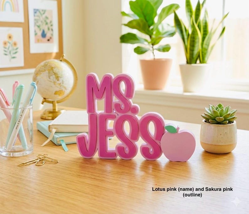Puede incluir: Un letrero de escritorio personalizado con el nombre MS. JESS en letras rosas loto en relieve y negrita con un contorno rosa sakura, acompa&ntilde;ado de un peque&ntilde;o icono con forma de manzana rosa claro unido al lado derecho del nombre.