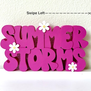 May include: A vibrant magenta sign with the words "SUMMER STORMS" in large, rounded letters. White daisy accents are placed above and below the text. The phrase "Swipe Left" is above the sign.
