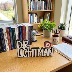 Puede incluir: Un letrero de escritorio en blanco y negro que dice DR. LICHTMAN con un logotipo circular de Alabama Crimson Tide en el lado derecho. El letrero se encuentra sobre un escritorio de madera junto a libros, una taza blanca con texto UA y plantas.