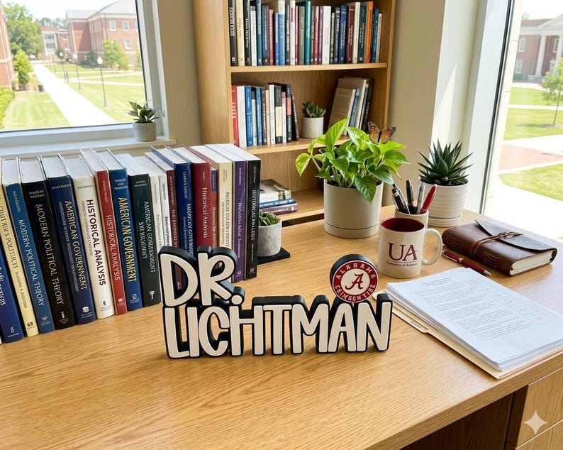 Puede incluir: Un letrero de escritorio en blanco y negro que dice DR. LICHTMAN con un logotipo circular de Alabama Crimson Tide en el lado derecho. El letrero se encuentra sobre un escritorio de madera junto a libros, una taza blanca con texto UA y plantas.