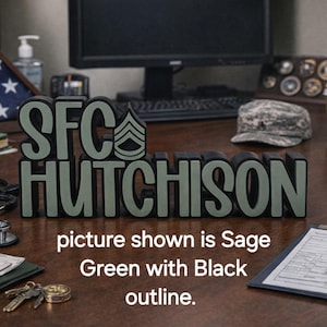 May include: Sage green and black SFC HUTCHISON desk sign. The sign is displayed on a desk with a military theme, including a folded American flag, a military cap, and a stethoscope. The text "picture shown is Sage Green with Black outline" is also visible.