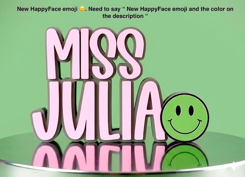 Puede incluir: Un letrero tridimensional con el texto MISS JULIA en letras de color rosa claro con bordes gris oscuro, acompa&ntilde;ado de un emoji de cara feliz de color verde brillante con ojos y boca negros, todo sobre una superficie reflectante.
