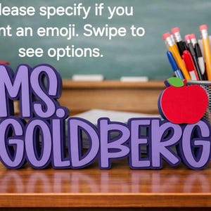 Puede incluir: Un letrero 3D morado que dice MS. GOLDBERG con un icono de manzana roja y una hoja verde. El texto dice: Please specify if you want an emoji. Swipe to see options. Una pila de libros y un portal&aacute;pices de malla met&aacute;lica con varios bol&iacute;grafos.