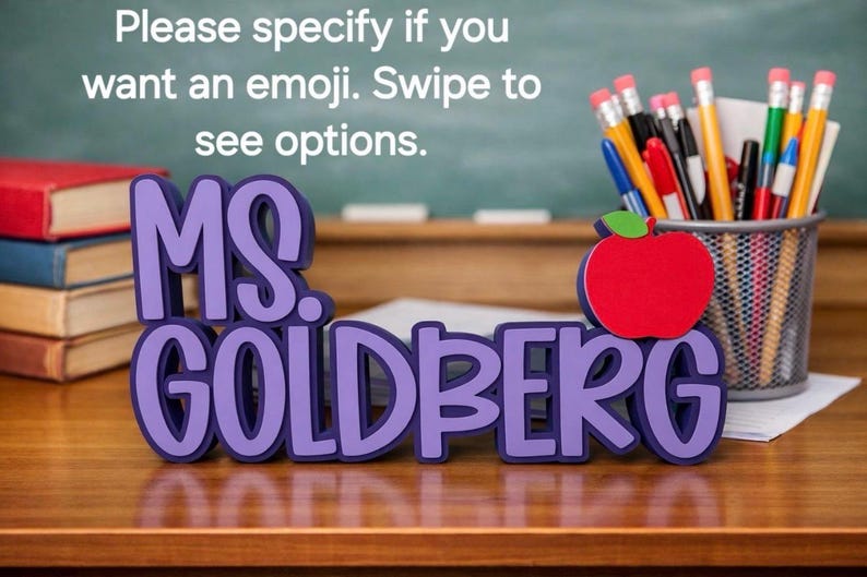 Puede incluir: Un letrero 3D morado que dice MS. GOLDBERG con un icono de manzana roja y una hoja verde. El texto dice: Please specify if you want an emoji. Swipe to see options. Una pila de libros y un portal&aacute;pices de malla met&aacute;lica con varios bol&iacute;grafos.