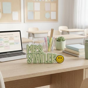 Puede incluir: Un letrero de escritorio 3D en color verde claro que muestra el texto MRS. STATLER en una fuente redondeada, acompa&ntilde;ado de un peque&ntilde;o icono de cara sonriente amarilla. El letrero est&aacute; sobre un escritorio de madera junto a un port&aacute;til.