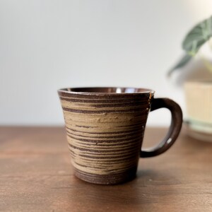 May include: A brown and tan striped ceramic mug with a handle. The mug is sitting on a wooden surface.