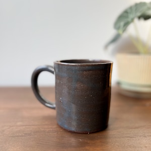 May include: A dark brown ceramic mug with a rounded handle. The mug has a textured surface and a slightly irregular shape, suggesting it is handmade. The mug is sitting on a wooden surface.