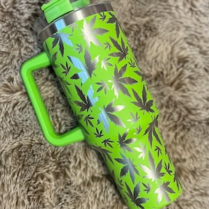 May include: A green stainless steel travel mug with a black and silver marijuana leaf pattern. The mug has a handle and a lid.