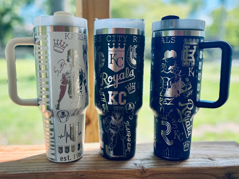 May include: Three stainless steel travel mugs with black and silver designs featuring the Kansas City Royals logo and team graphics. The mugs have handles and lids.