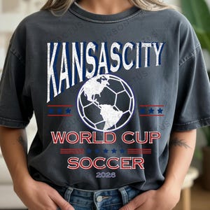Puede incluir: Camiseta gris oscuro con la inscripción "KANSAS CITY" en blanco y azul, y un gráfico de balón de fútbol que representa la Tierra. Debajo, se lee "WORLD CUP SOCCER 2026" en rojo y blanco. El diseño incluye estrellas y rayas.