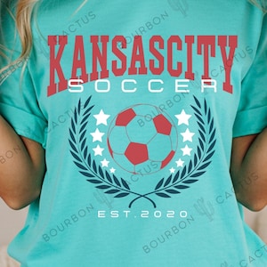 Puede incluir: Camiseta turquesa con la inscripción "KANSAS CITY SOCCER" en rojo. El diseño incluye un balón de fútbol rojo y blanco, estrellas blancas y una corona de laurel azul oscuro. El texto "EST. 2020" está debajo de la corona.