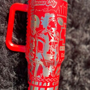 May include: Red stainless steel travel mug with a black handle. The mug has a silver design featuring a football player, the words "Kansas City Chiefs" and a skyline.