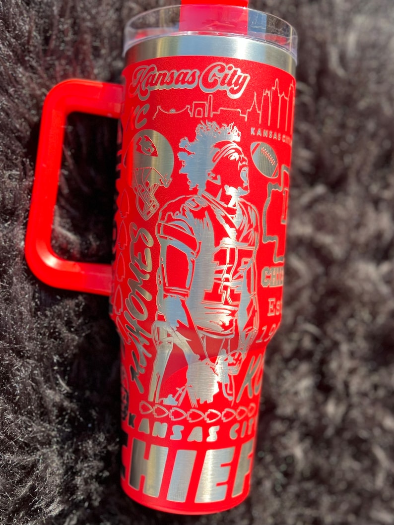 May include: Red stainless steel travel mug with a black handle. The mug has a silver design featuring a football player, the words "Kansas City Chiefs" and a skyline.
