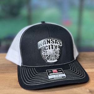 May include: Black and white trucker hat with a patch that says "Kansas City Missouri". The hat has a black front panel, a white mesh back, and a curved brim. The Richardson 112 Trucker hat is adjustable.