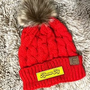 May include: Red cable knit beanie with a faux fur pom pom and a yellow patch that says "Kansas City" in a retro font.