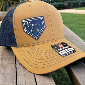May include: A brown and navy blue trucker hat with a gold and blue baseball-shaped patch that says "Kansas City". The hat has a Richardson 112 adjustable strap.