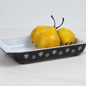 May include: A black baking dish with a white snowflake pattern around the rim. Three yellow pears are inside the dish.
