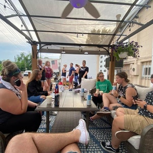 Puede incluir: Un grupo de personas sentadas alrededor de una mesa en un patio, algunas están bebiendo de latas y otras están en sus teléfonos. Hay un ventilador de techo blanco sobre la mesa.