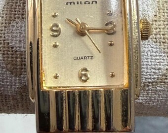 May include: A gold-toned Milan quartz watch with a rectangular face. The watch face is a light champagne color with black numerals and hands. The watch band has a ribbed design.