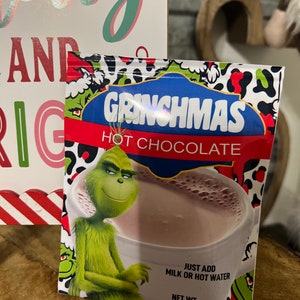 May include: A green Grinch character wearing a red and white striped hat is featured on a packet of Grinchmas hot chocolate mix. The packet is red, white and green with a black leopard print design. The text on the packet reads "Grinchmas Hot Chocolate" and "Just add milk or hot water. Net Wt. 0.73oz."