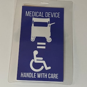 May include: A white plastic badge holder with a blue card inside. The card has a white silhouette of a medical cart with the text "MEDICAL DEVICE" above it and a wheelchair with the text "HANDLE WITH CARE" below it.