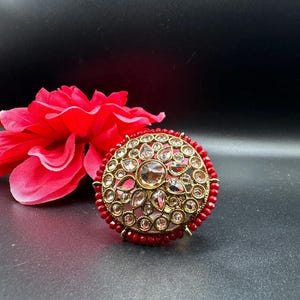 Peut inclure: Une bague dorée avec une grosse pierre centrale transparente et de plus petites pierres transparentes autour. La bague est entourée de petites perles rouges. Une fleur rouge est en arrière-plan sur une surface noire.