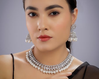 May include: A silver and white bridal jewellery set with a necklace, earrings, and a tikka. The necklace features a layered design with white pearls and clear stones. The earrings are dangle style with a similar design to the necklace. The tikka is a traditional Indian headpiece with a pearl and stone design.