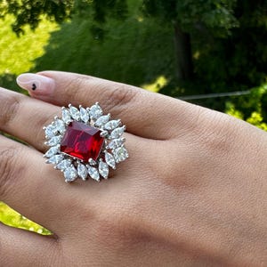 May include: A silver ring featuring a large, square, red gemstone surrounded by smaller, clear gemstones.
