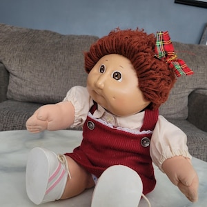 May include: A Cabbage Patch Kid doll with brown yarn hair, a red and white checked bow, and a red overall dress. The doll is sitting on a white surface.