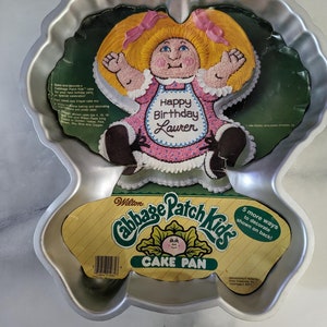 May include: A silver metal cake pan shaped like a Cabbage Patch Kid doll. The pan has a yellow label with the text "Wilton Cabbage Patch Kids Cake Pan" and a picture of a Cabbage Patch Kid doll. The text "Happy Birthday Lauren" is printed on the doll's shirt.