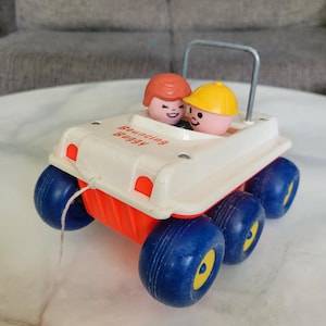 Puede incluir: Un coche de juguete blanco y rojo con ruedas azules y una cuerda para tirar. El coche tiene un letrero que dice "Bouncing Buggy" y dos figuras con caras sonrientes están sentadas dentro.