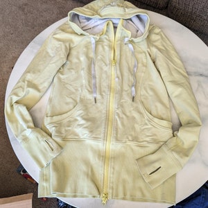 May include: A light yellow hooded sweatshirt with a yellow zipper and two pockets. The sweatshirt has a ribbed hem and cuffs.