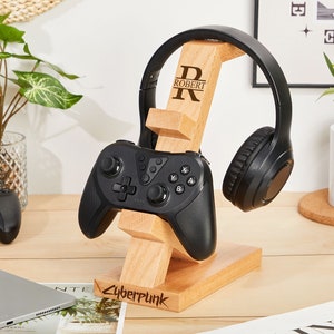May include: A wooden stand with a personalized inscription "R ROBERT" holds a black gaming headset and a black video game controller. The stand is engraved with the word "Cyberpunk".