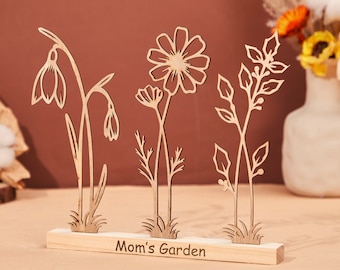May include: Wooden cutout of a flower arrangement with the text "Mom's Garden" on a wooden base. The flowers include a daisy, a snowdrop, and a sprig of leaves.